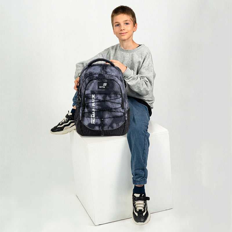 Model obok plecaka K26-772M-3 prezentujący jego proporcje i uchwyt na deskorolkę.